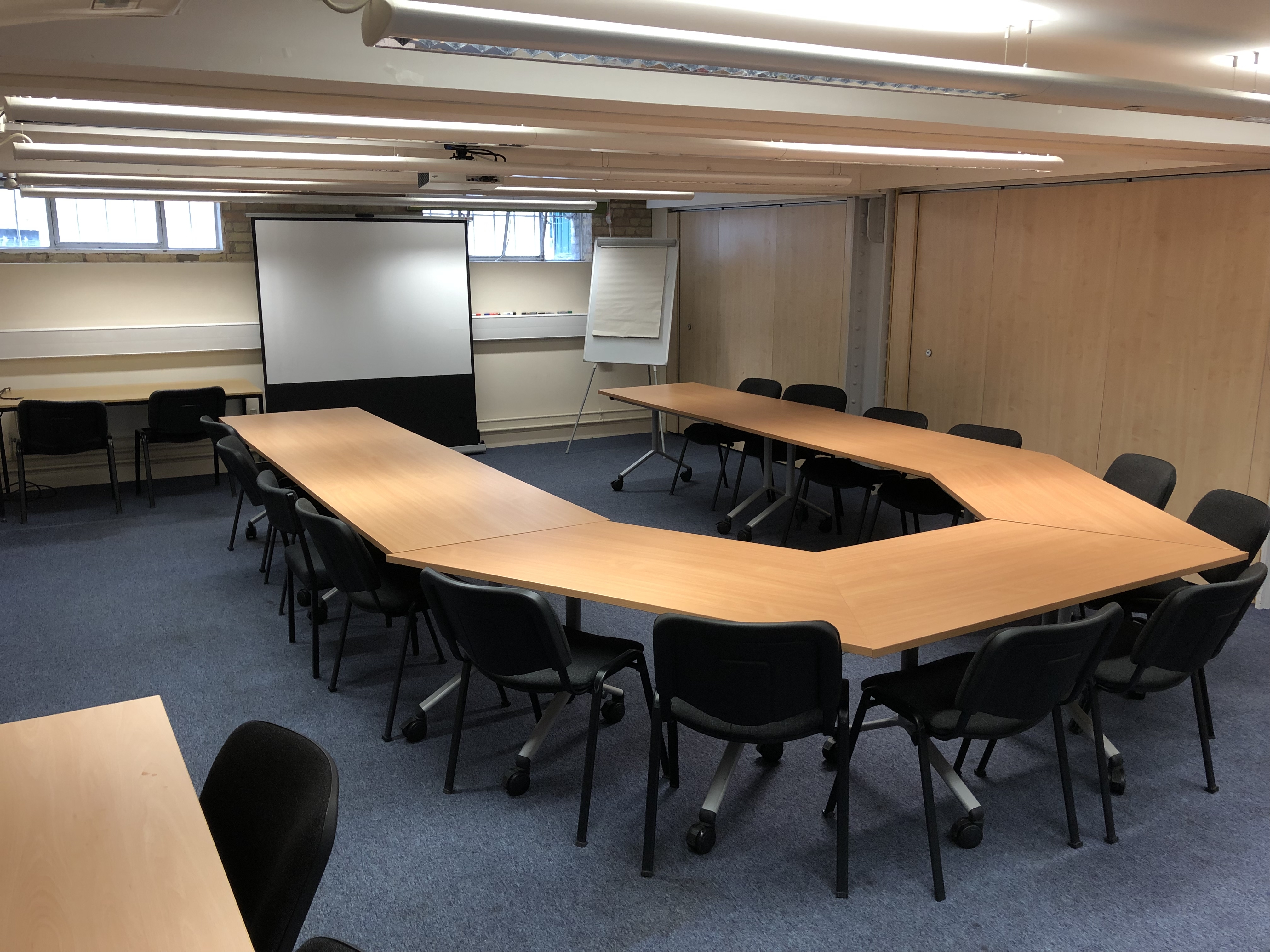 Mill Lane Seminar Room Photograph showing the standard layout of the Mill Lane Seminar Room - a room with fourteen chairs around a large boardroom table, with a projector screen on the end wall.